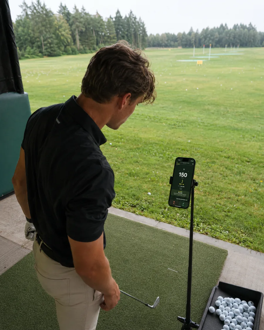 A golfer at a driving range bay with a phone on a stand showing the next shot call: 150 yards, draw.